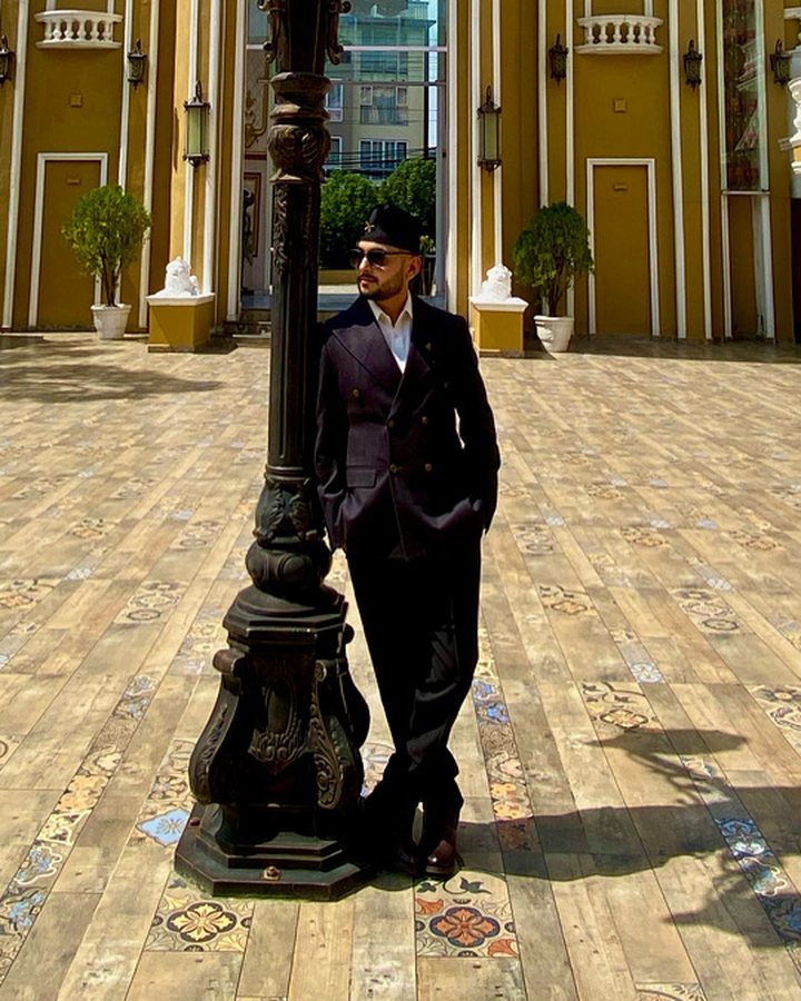 Sushant Mishra in a dark double-breasted suit and dhaka topi, standing beside an ornate cast-iron clock lamp-post in a paved courtyard in front of an ochre building.