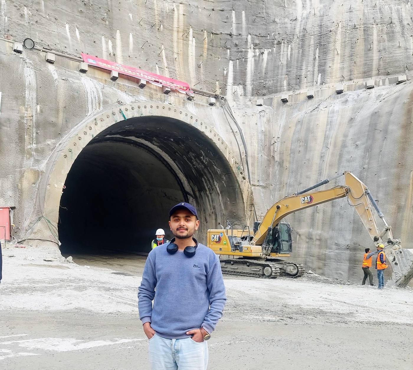 Sushant Mishra standing in front of the Siddhababa tunnel portal: a rock-bolted cut face, the concrete-lined tunnel bore behind him, a CAT excavator working the spoil to his right, and two workers in orange vests at the foot of the slope.
